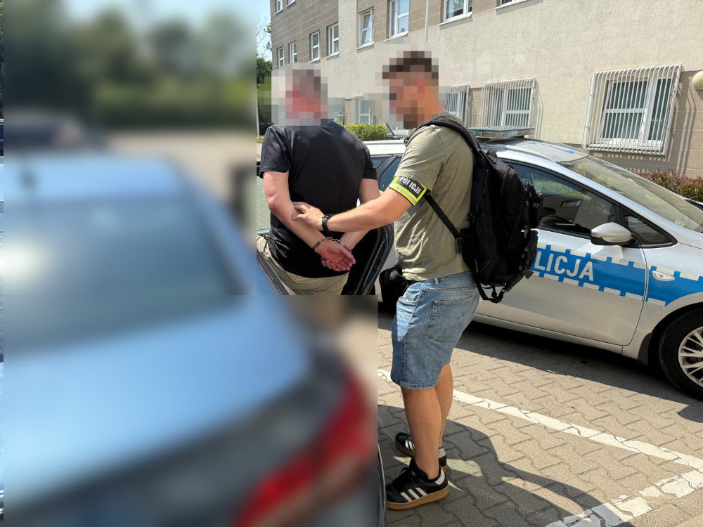 Mężczyzna w kajdankach jest zatrzymywany przez funkcjonariusza policji obok radiowozu na parkingu.