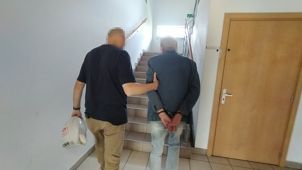 Mężczyzna w kajdankach prowadzony przez policjanta po schodach w budynku administracyjnym.