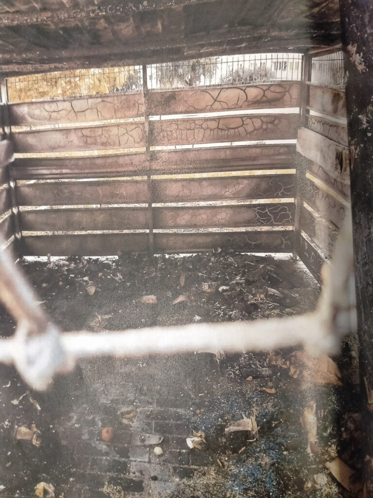 Pusty boks z drewnianymi ścianami, z widocznymi śladami zniszczeń i zanieczyszczeniami na podłodze.