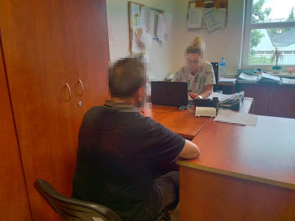 Mężczyzna siedzący na krześle rozmawia z kobietą przy biurku w biurze, obok laptopa i dokumentów.