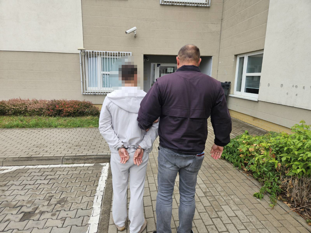 Mężczyzna w szarym dresie prowadzony przez policjanta na chodniku przed budynkiem mieszkalnym.