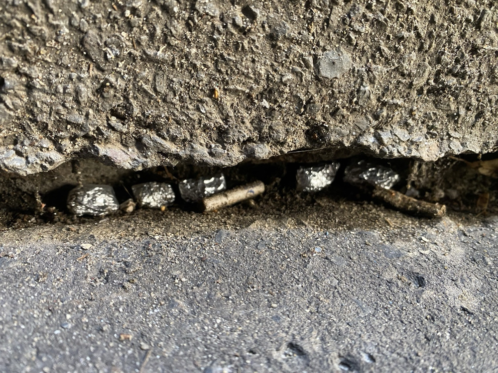 Skrzynka z metalowymi przedmiotami oraz odpadami umieszczona w szczelinie między betonowymi płytami.