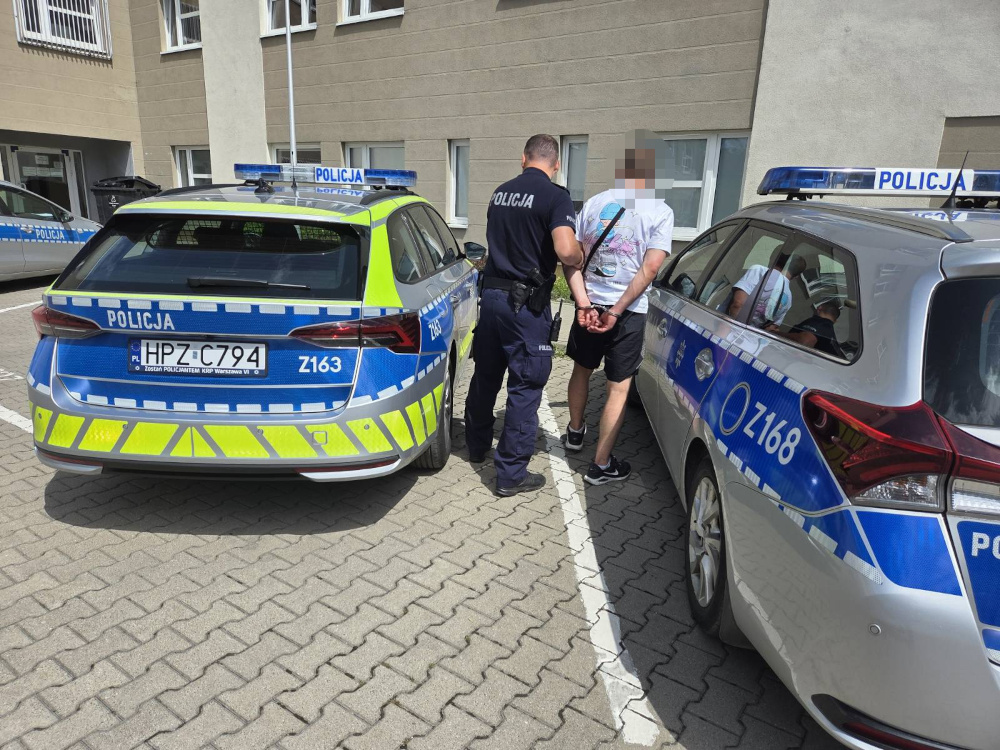 Mężczyzna w koszulce z rękami związanymi z tyłu stoi obok policyjnego radiowozu na osiedlowym parkingu.