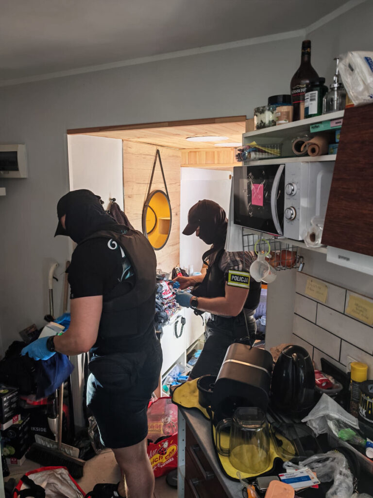 Funkcjonariusze policji przeszukują mieszkanie, wypełnione różnymi przedmiotami, w poszukiwaniu dowodów.