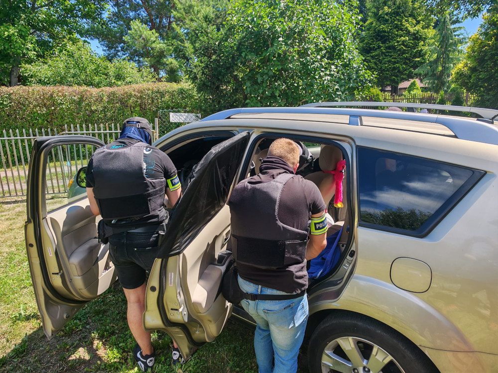 Dwaj policjanci w kamizelkach taktycznych przeszukują wnętrze samochodu w otoczeniu zieleni.