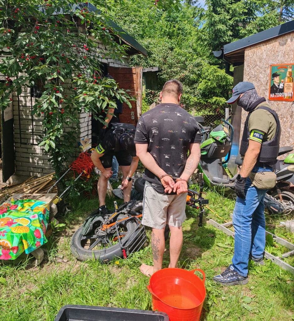 Mężczyzna z rękami związanymi z tyłu stoi na trawie obok dwóch policjantów, którzy przeszukują teren przy budynku.