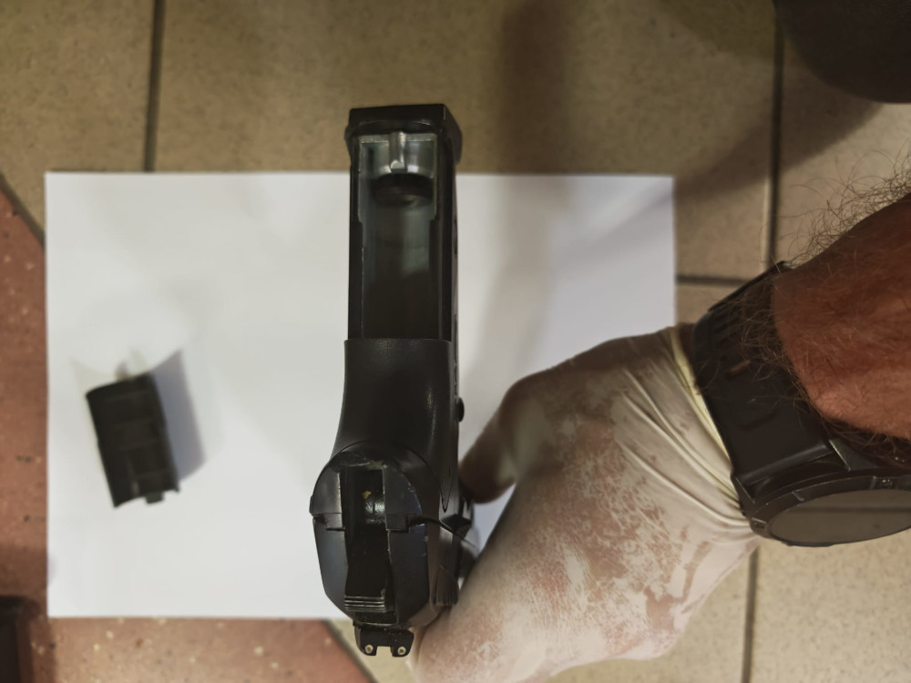 Pistolet leży na białym arkuszu papieru, obok znajduje się magazynki. Widać rękę w rękawiczce trzymającą broń.