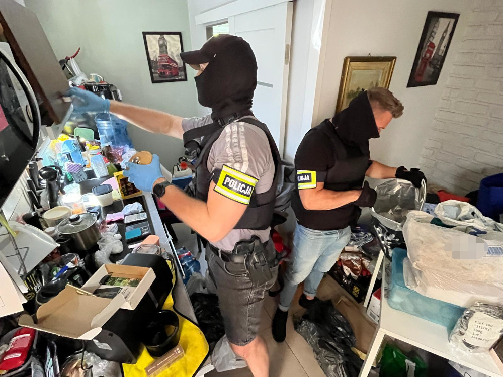 Dwóch policjantów w maskach przeszukuje zagracone pomieszczenie, przeszukując szafki i stoły.