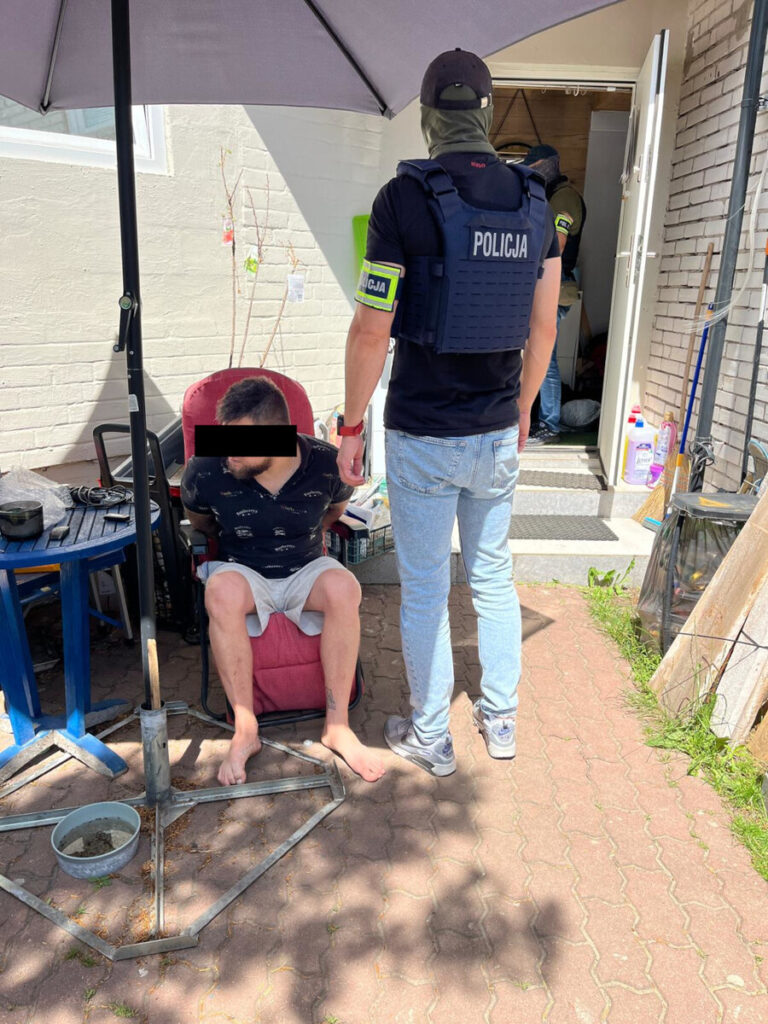 Mężczyzna w krótkich spodniach siedzi na krześle, podczas gdy funkcjonariusz policji w kamizelce taktycznej stoi obok.