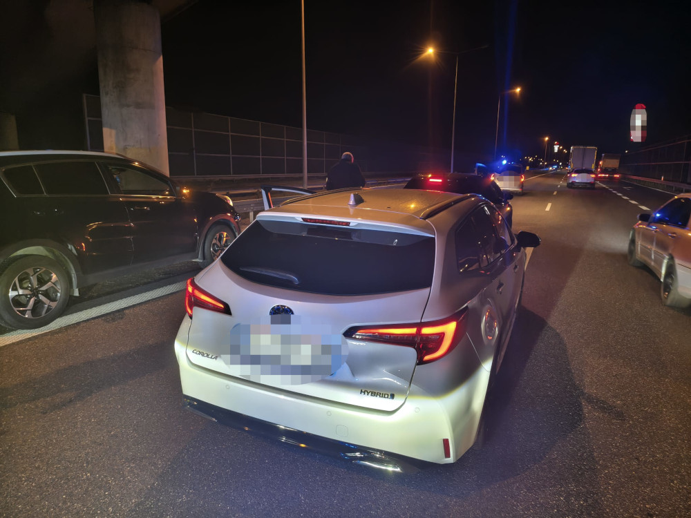Biały samochód marki Toyota zaparkowany na autostradzie w nocy, obok stojące inne pojazdy oraz radiowóz z włączonymi światłami.