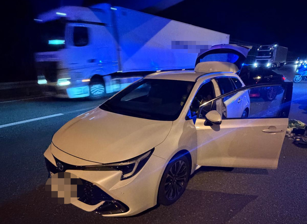 Biała Toyota z otwartymi drzwiami zatrzymana na autostradzie, obok przejeżdżający ciężarówka w nocy.