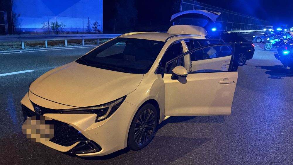 Biała Toyota z otwartymi drzwiami, zatrzymana na autostradzie w nocy, otoczona przez radiowozy policyjne.
