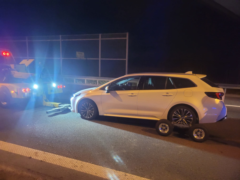 Biały samochód osobowy unieruchomiony na autostradzie, holowany przez pojazd służbowy z włączonymi światłami.