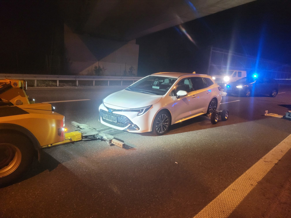 Biały samochód osobowy holowany przez pojazd pomocy drogowej na autostradzie w nocy, w tle niebieskie światła radiowozu.