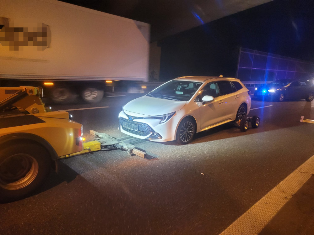 Biały samochód marki Toyota holowany przez lawetę na autostradzie w nocy, obok przejeżdżające ciężarówki.