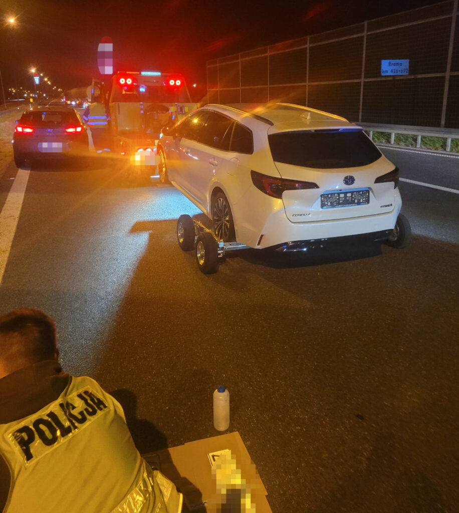 Policjant w kamizelce odblaskowej bada skradzioną Toyotę na autostradzie, obok stoi laweta z sygnalizacją.