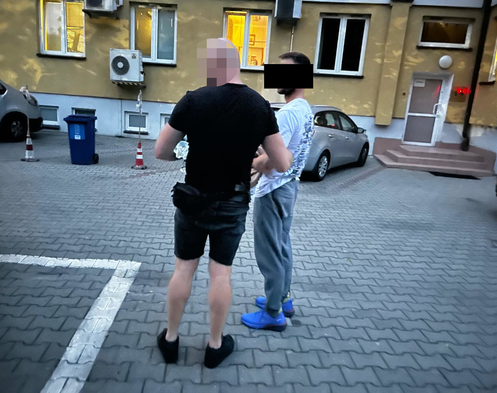 Mężczyzna w czarnym t-shircie rozmawia z drugim mężczyzną w jasnej koszulce na terenie podwórka budynku mieszkalnego.