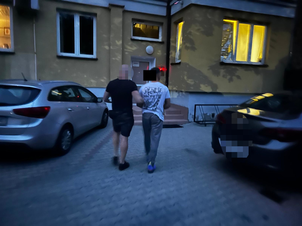 Mężczyzna w szarych spodniach prowadzony przez funkcjonariusza w kierunku budynku z podświetloną wejściem.