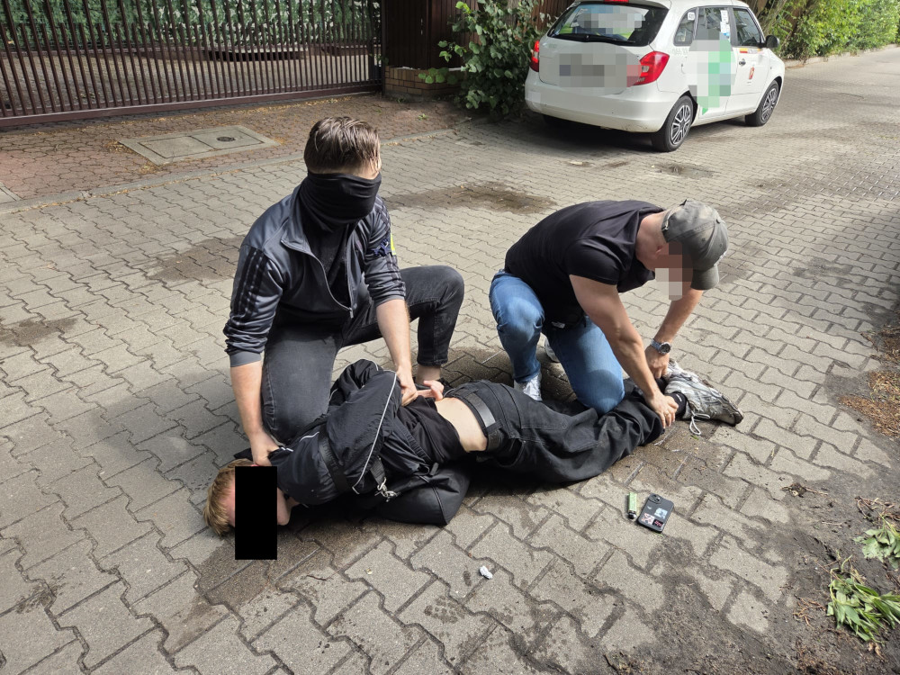Dwaj mężczyźni w ciemnych ubraniach zatrzymują osobę leżącą na chodniku, w tle biały samochód.