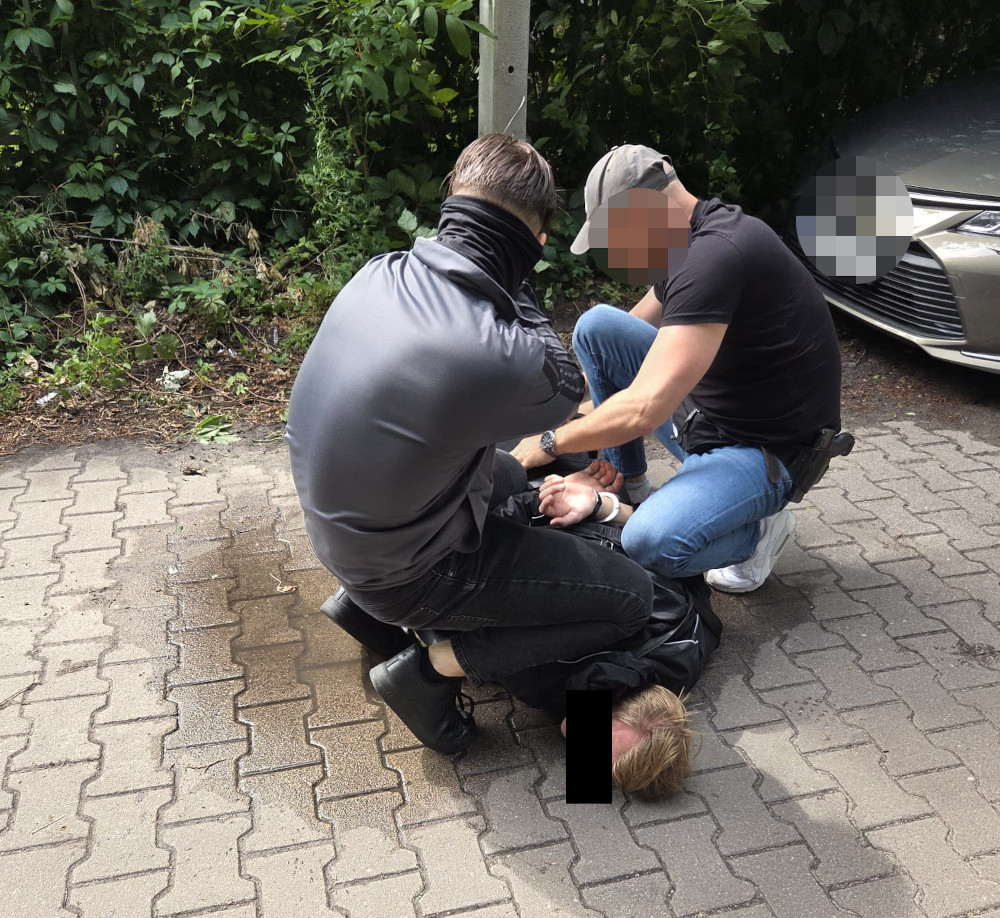 Dwaj mężczyźni w odzieży cywilnej zatrzymują osobę na chodniku, przy metalowym słupku w otoczeniu zieleni.