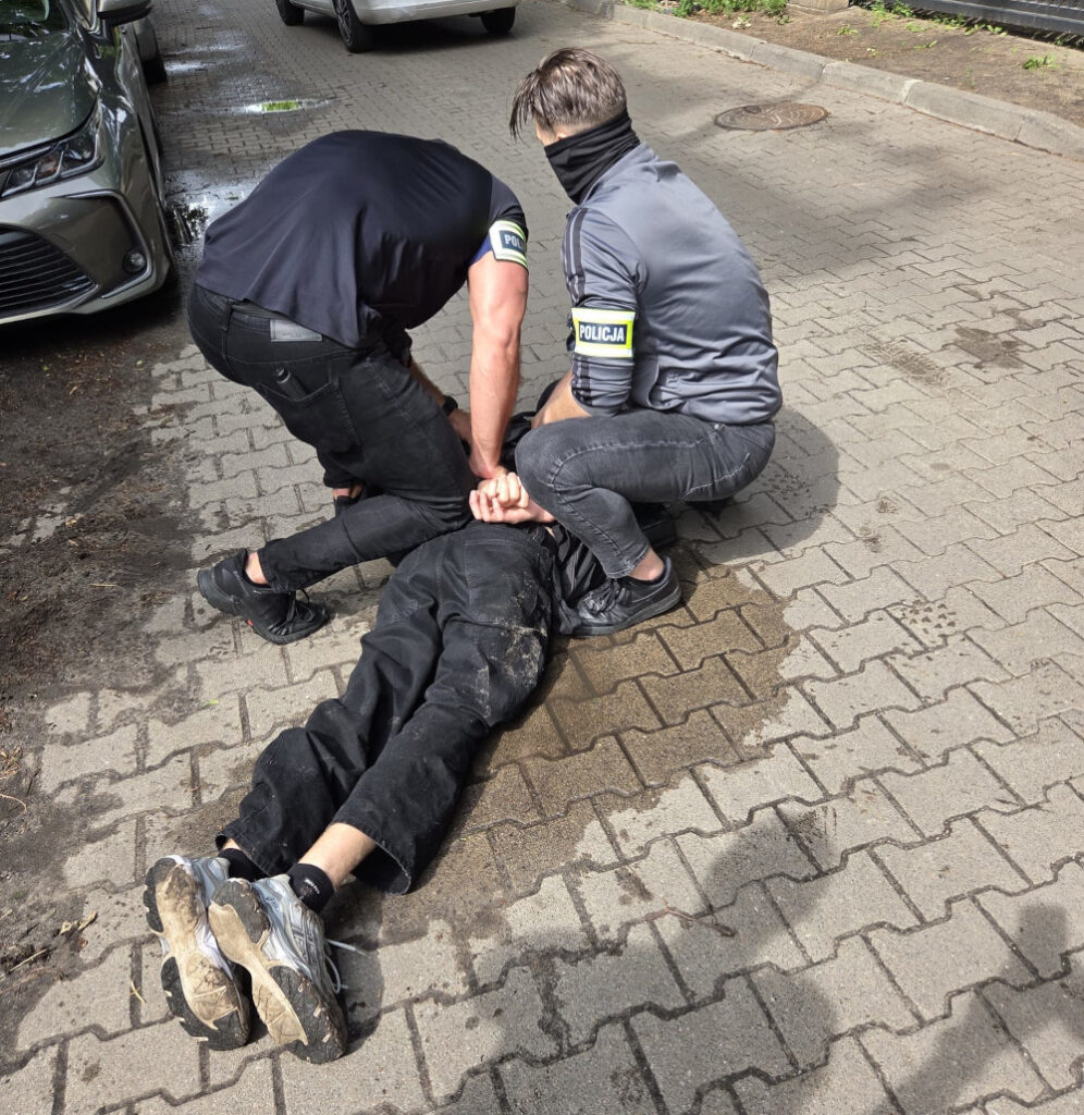 Dwóch policjantów trzyma mężczyznę leżącego na ziemi w brudnych ubraniach na wyłożonym kostką chodniku.