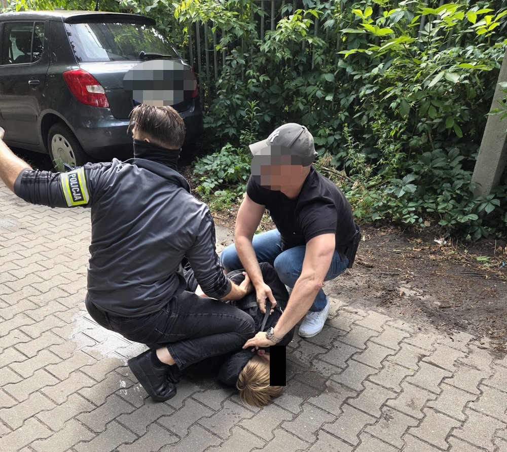 Policjant w mundurze unosi rękę, podczas gdy dwóch mężczyzn obezwładnia osobę leżącą na ziemi w zaroślach.