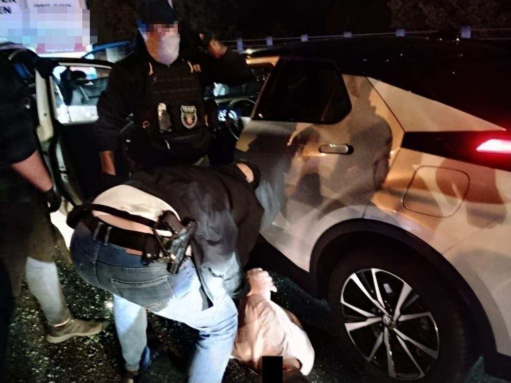 Funkcjonariusze policji zatrzymują mężczyznę w białej odzieży przy czarnym samochodzie na oświetlonej ulicy.