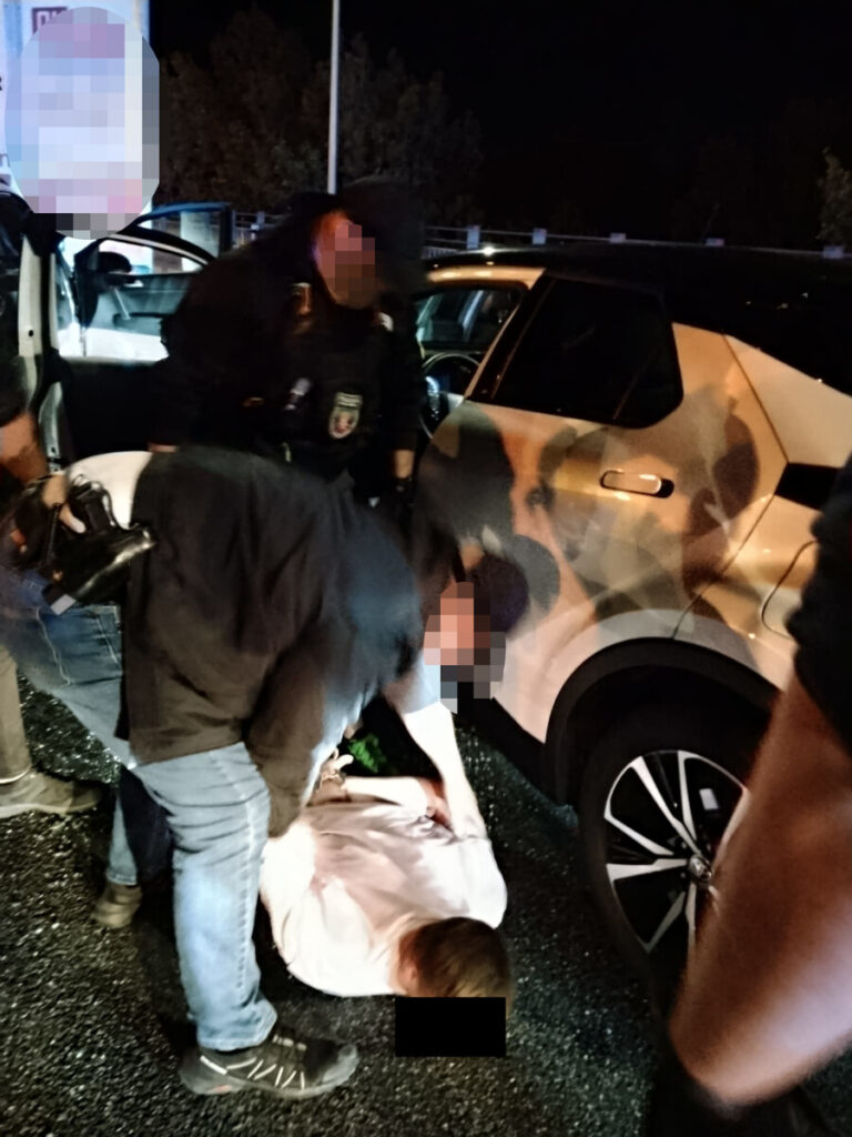 Funkcjonariusze policji zatrzymują mężczyznę leżącego na ziemi obok samochodu w nocy.