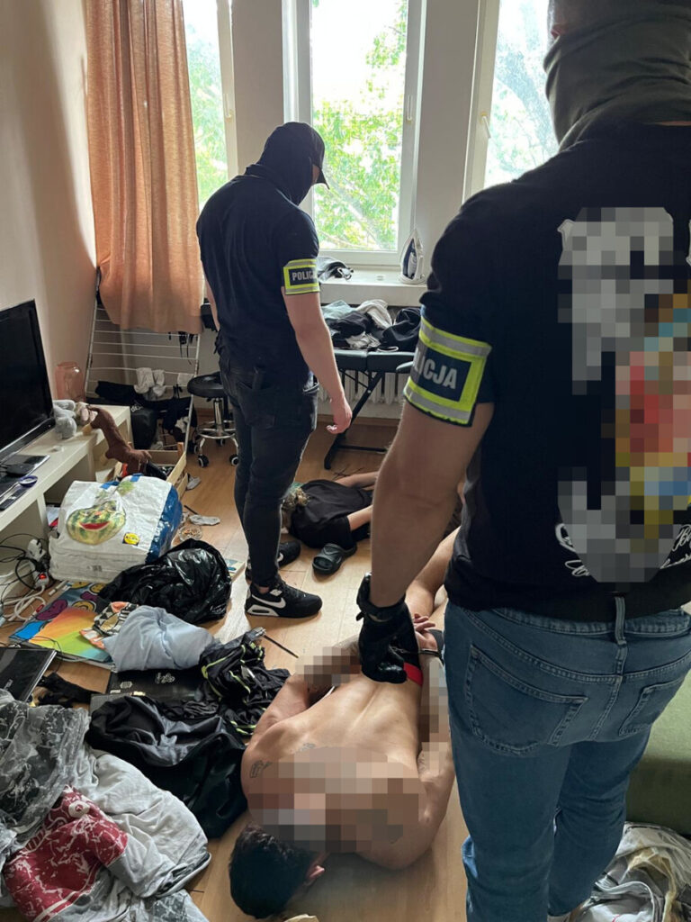 Policjanci w maskach przeszukują mieszkanie, gdzie leży mężczyzna na podłodze w bieliźnie, a wokół rozrzucone ubrania.