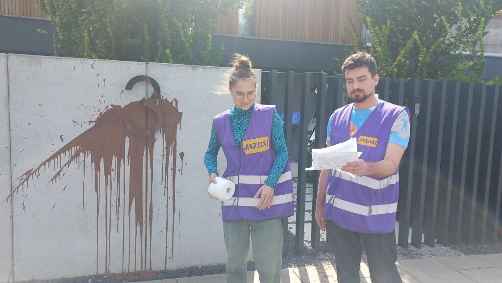 Mężczyzna i kobieta w fioletowych kamizelkach z napisem "JAZDA!" trzymają dokumenty, stojąc przed betonowym ogrodzeniem z plamami.