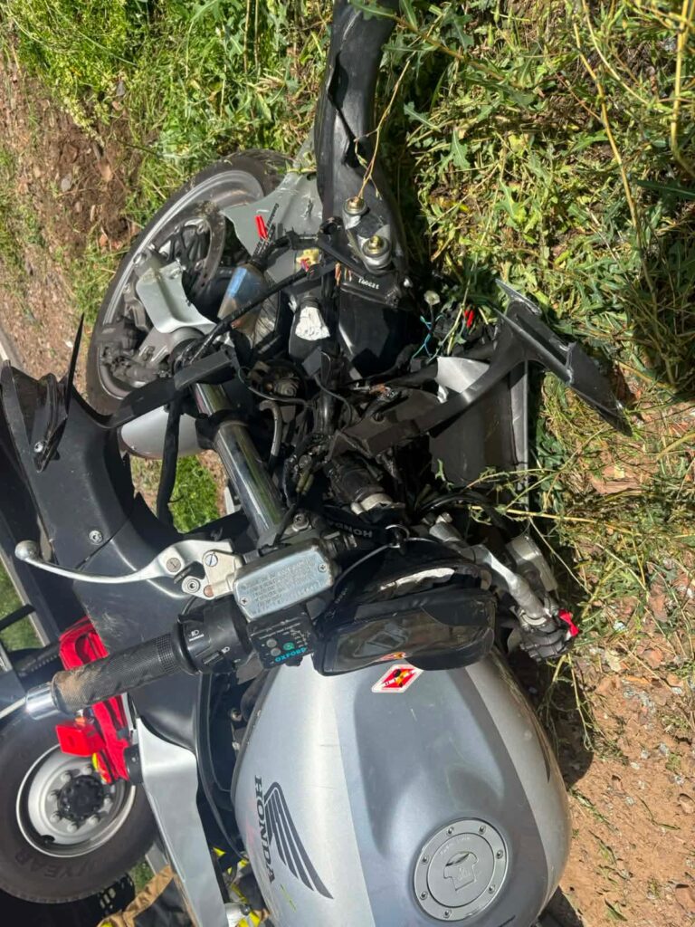 Zniszczony motocykl leżący na ziemi wśród wysokiej trawy, z widocznymi uszkodzeniami kierownicy i silnika.