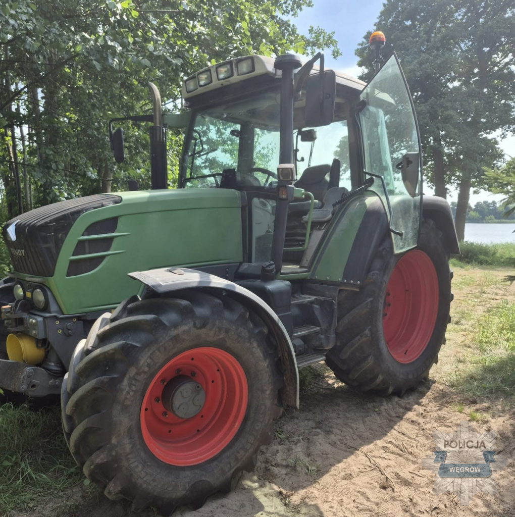 Traktor z zieloną kabiną i czerwonymi felgami stoi na piaszczystym terenie w otoczeniu drzew, w pobliżu wody.