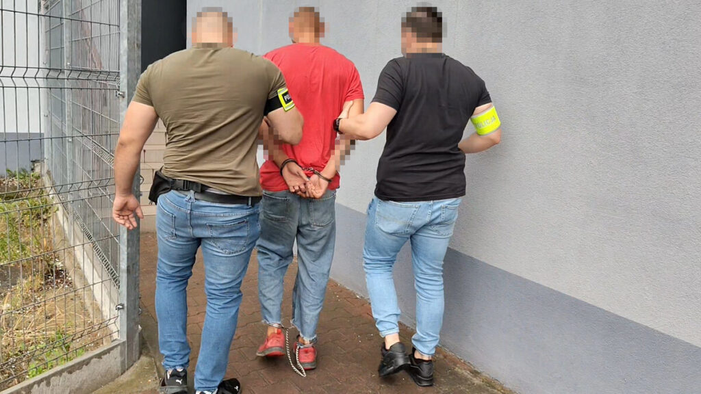Trzech funkcjonariuszy policji prowadzi aresztowanego mężczyznę z rękami związanymi z tyłu w miejskim otoczeniu.