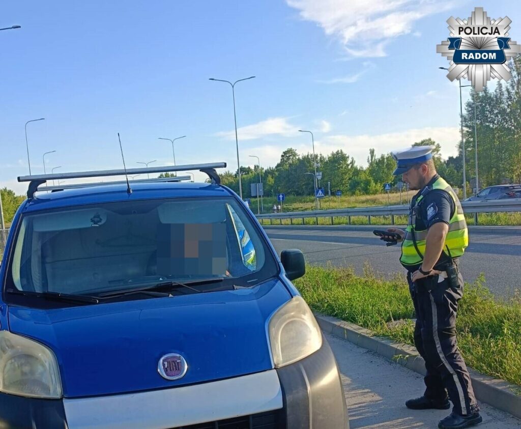 Policjant w kamizelce odblaskowej kontroluje niebieski pojazd dostawczy na poboczu drogi w słoneczny dzień.