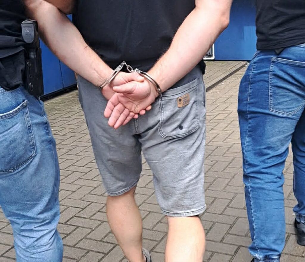 Mężczyzna w szortach z rękami skuty w kajdankach prowadzony przez dwóch policjantów po chodniku.