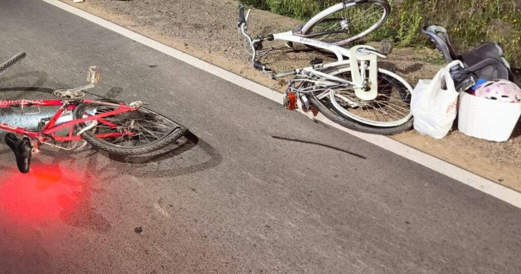 Na drodze leży przewrócony czerwony rower, obok znajduje się biały rower oraz torba z różnymi przedmiotami.