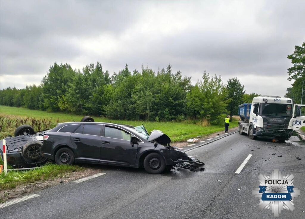 Czarny samochód osobowy leży na boku obok uszkodzonego pojazdu ciężarowego na drodze otoczonej drzewami.