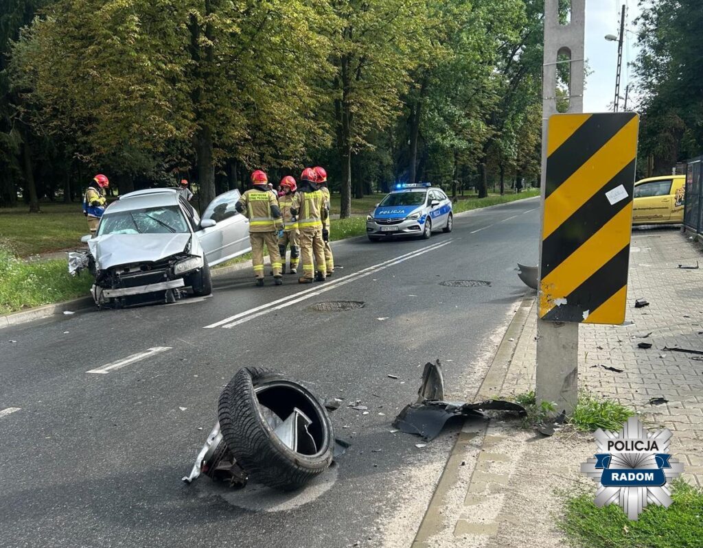 Grupa strażaków w hełmach bada uszkodzony samochód osobowy na poboczu drogi, obok stoi policyjny radiowóz.