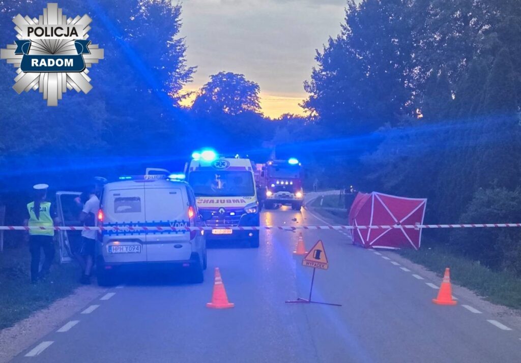 Policjanci zabezpieczają miejsce wypadku drogowego, w tle widoczne pojazdy służb ratunkowych.