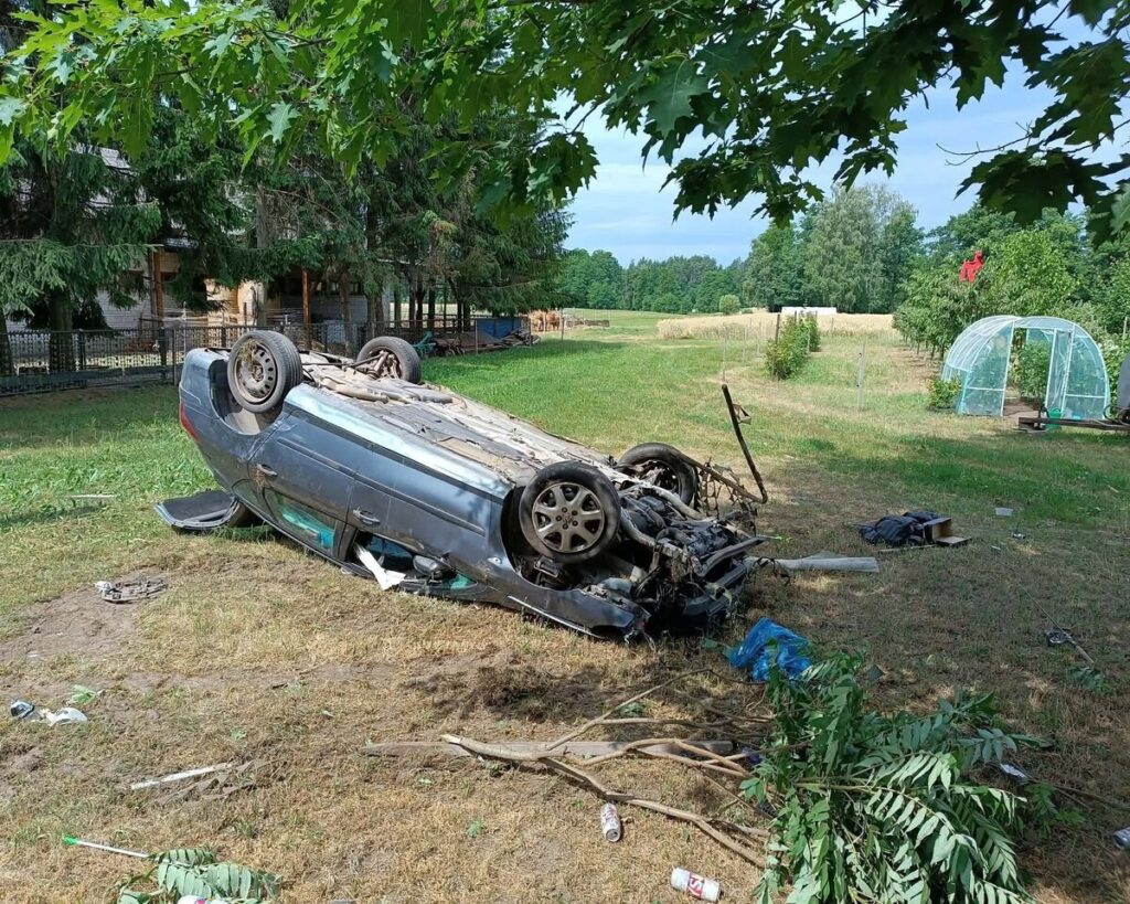 Przewrócony samochód leży na trawie w wiejskim otoczeniu, w pobliżu drzew i szklanej szklarni.