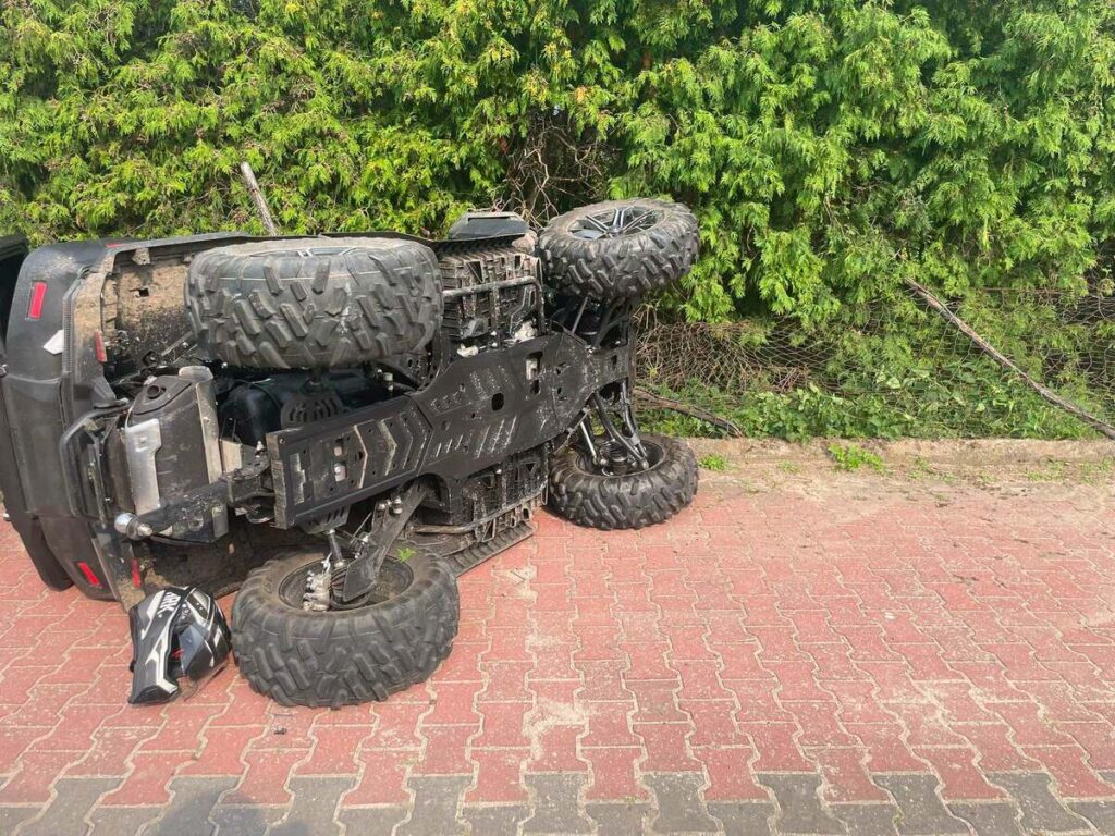 Przewrócony quad leży na drodze, obok niego kask oraz zieleń w tle.