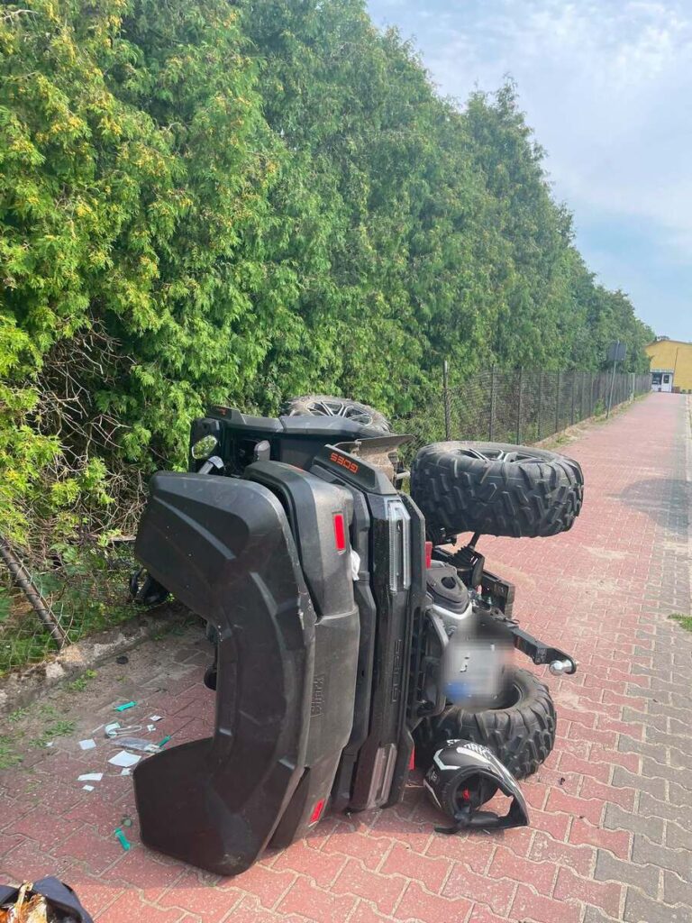 Quadowy pojazd leży na boku na chodniku, otoczony gęstą zielenią i ogrodzeniem.