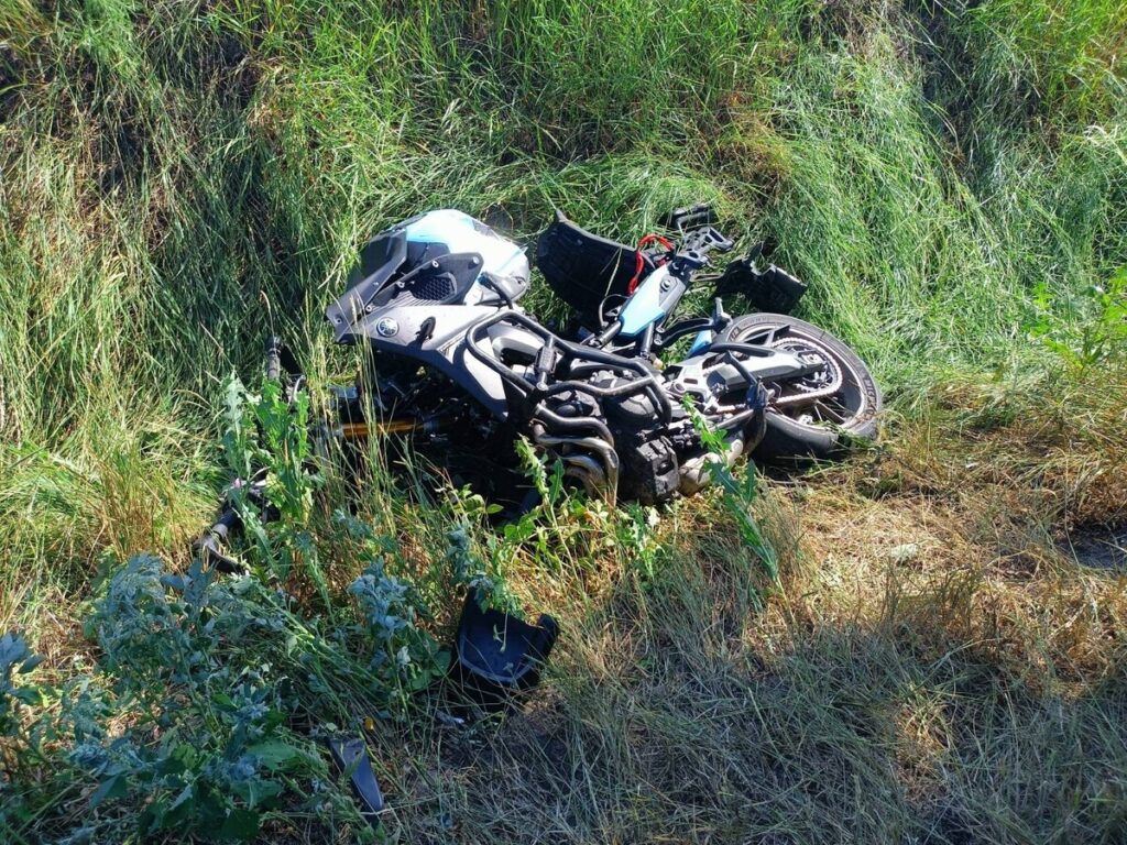 Motocykl leży przewrócony w wysokiej trawie, otoczony zielenią i dzikimi roślinami.