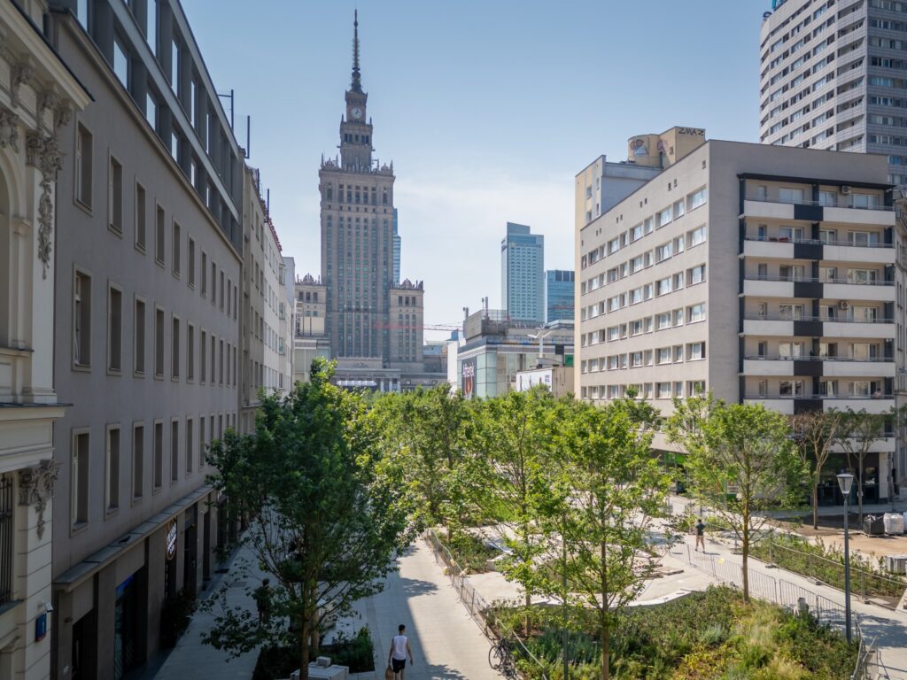 Wysoki budynek z wieżą, będący przykładem architektury socrealistycznej, widoczny w tle nowoczesnej przestrzeni miejskiej z drzewami.