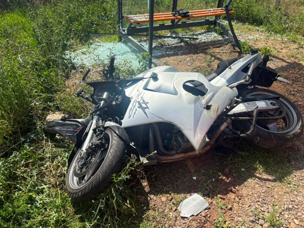 Przewrócony motocykl leży na trawie obok ławki w otoczeniu wysokiej trawy i fragmentów szkła.