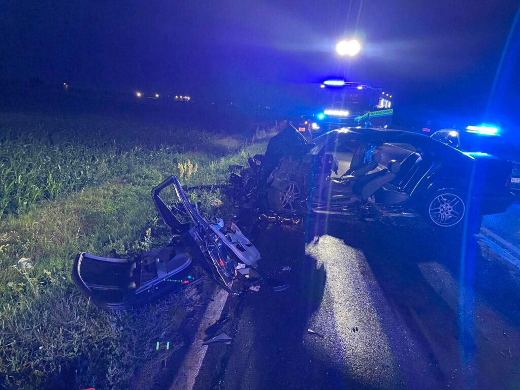 Zniszczony samochód osobowy leży na poboczu drogi, obok widać uszkodzone elementy pojazdu, a w tle migają niebieskie światła radiowozów.