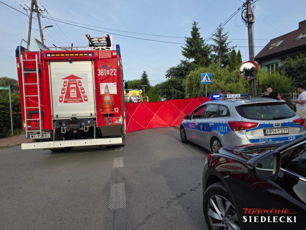 Na drodze stoi wóz strażacki oraz radiowóz policyjny, a za nimi rozwinięta czerwona taśma blokująca dostęp do miejsca zdarzenia.
