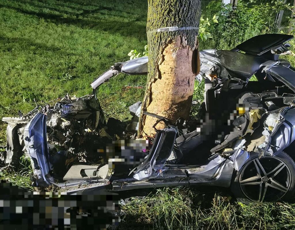Zniszczony samochód osobowy wbił się w drzewo, leżący na trawie w otoczeniu zieleni.