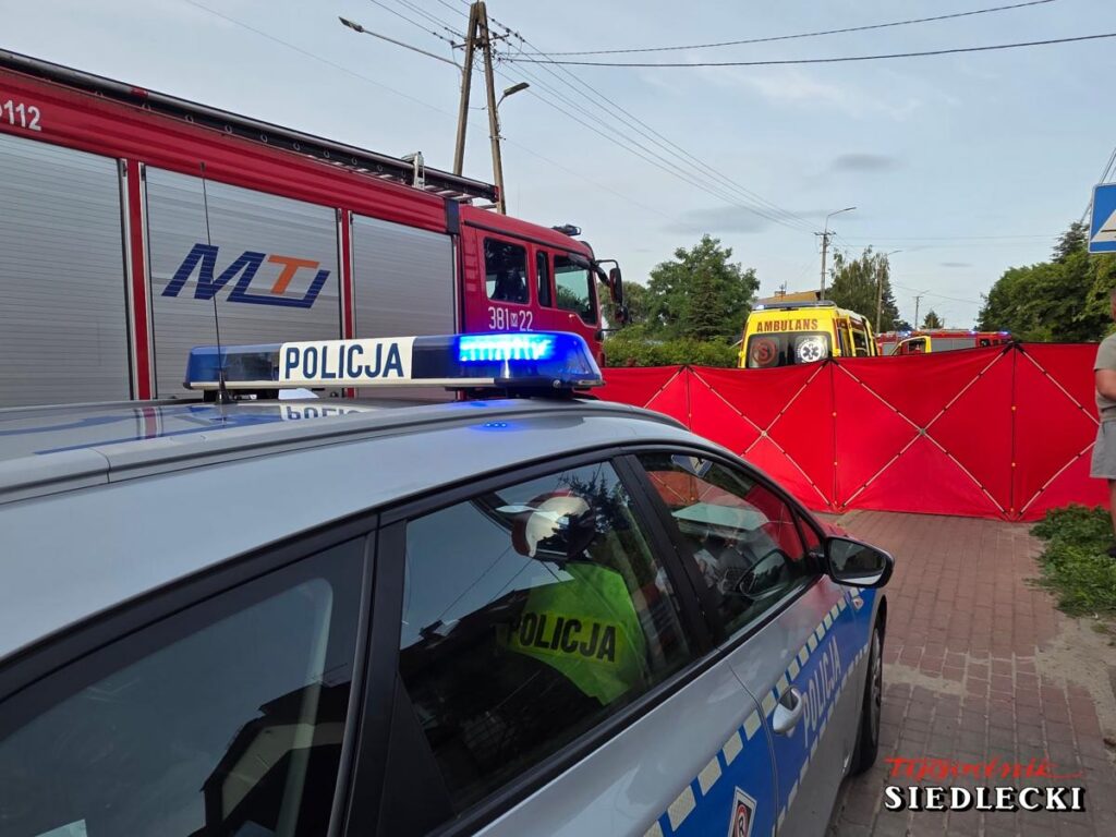 Policyjny radiowóz z włączonymi sygnałami świetlnymi stoi obok czerwonego samochodu strażackiego, a w tle widoczne są karetki pogotowia oraz ogrodzenie zabezpieczające miejsce zdarzenia.