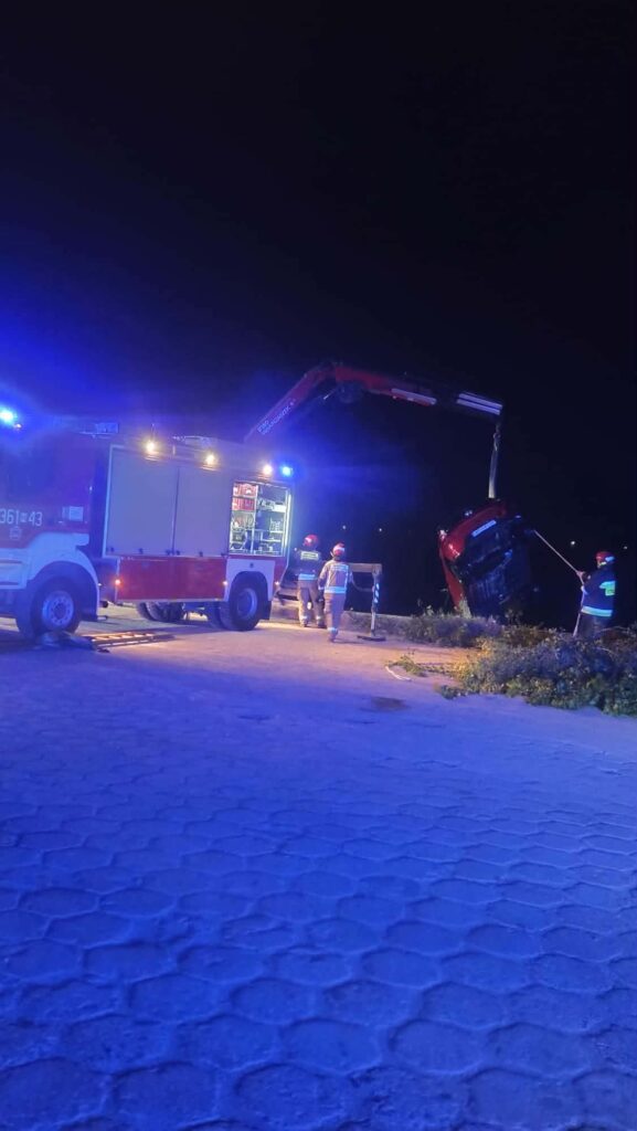 Strażacy używają wozu strażackiego do podnoszenia przewróconego samochodu na nocnej drodze.